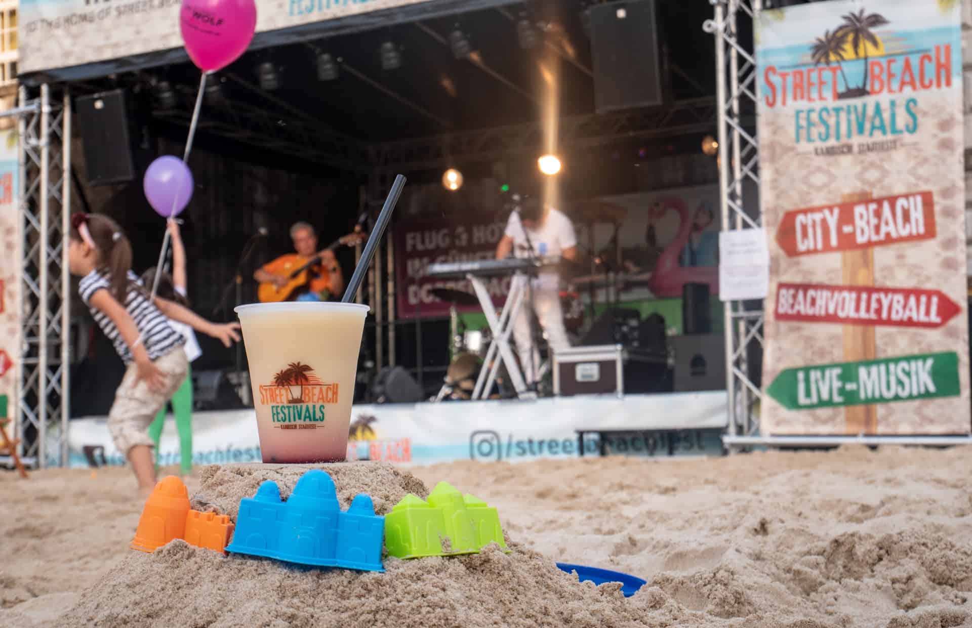Auf einer Sandfläche stehen auf einem kleinen Sandhügel Sandförmchen und ein Getränkebecher mit Logo des Streetfood-Beach-Festivals. Im Hintergrund ist eine Bühne zu sehen. Auf einer Sandfläche stehen auf einem kleinen Sandhügel Sandförmchen und ein Getränkebecher als Symbolbild für Gourmet Festivals